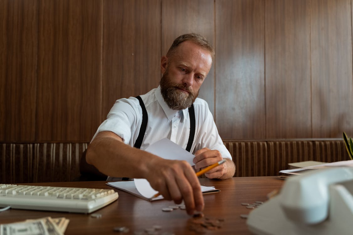 A pensive financial analyst with a beard, counting coins and examining paperwork, highlighting the meticulous nature of cost analysis.