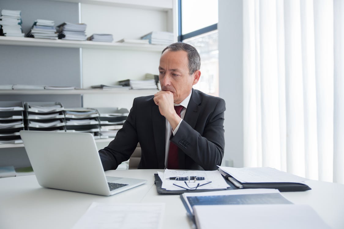 A contemplative executive analyzing data on a laptop in a modern office environment, with documents spread on the desk.