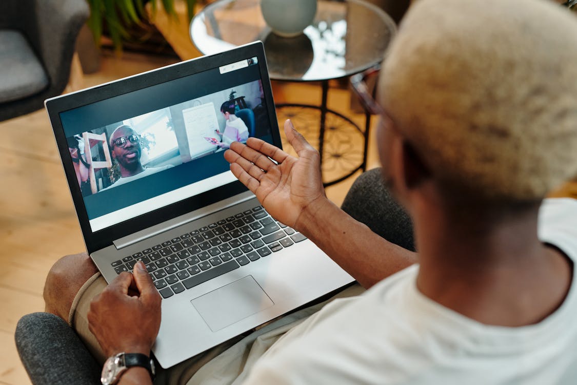 A person is seen from behind, gesturing with their hand while engaging in a video conference on a laptop. The laptop screen displays two other participants: one is smiling and wearing glasses, and the other appears to be focused on something outside the view. The setting suggests a casual, home environment.
