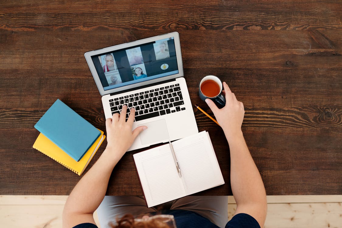 Top-down view of a person sitting at a wooden table with a laptop showing a multi-person video call. The individual is holding a cup of tea or coffee in one hand while navigating the laptop with the other. An open notebook with a pen in the center and two closed notebooks are also on the table, suggesting note-taking or multitasking during the meeting.