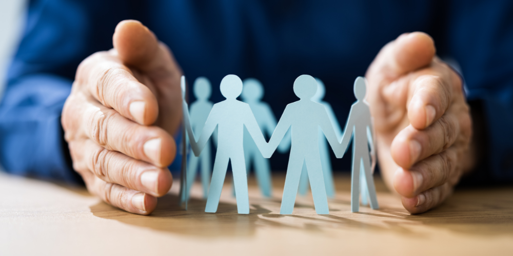 Hands carefully encircling a circle of paper cut-out figures on a table, depicting the concept of team unity and protection in the workplace.