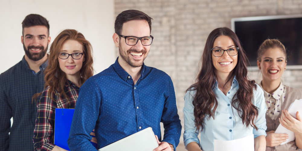 A group of five cheerful professionals with documents in hand, standing in an office setting, representing a team ready to collaborate and engage in the workplace.