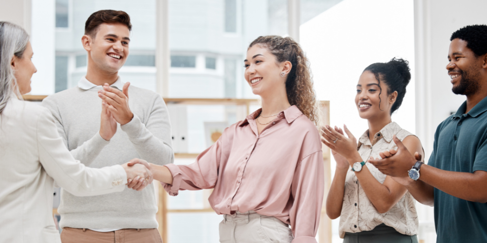 A group of diverse colleagues smiling and applauding as two people shake hands, representing a positive and inclusive employee welcoming
