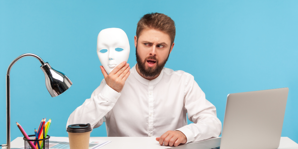A man sitting at a desk with a laptop, holding a plain white mask in one hand. There's a desk lamp, a coffee cup, and colorful pencils on the desk, against a blue background.