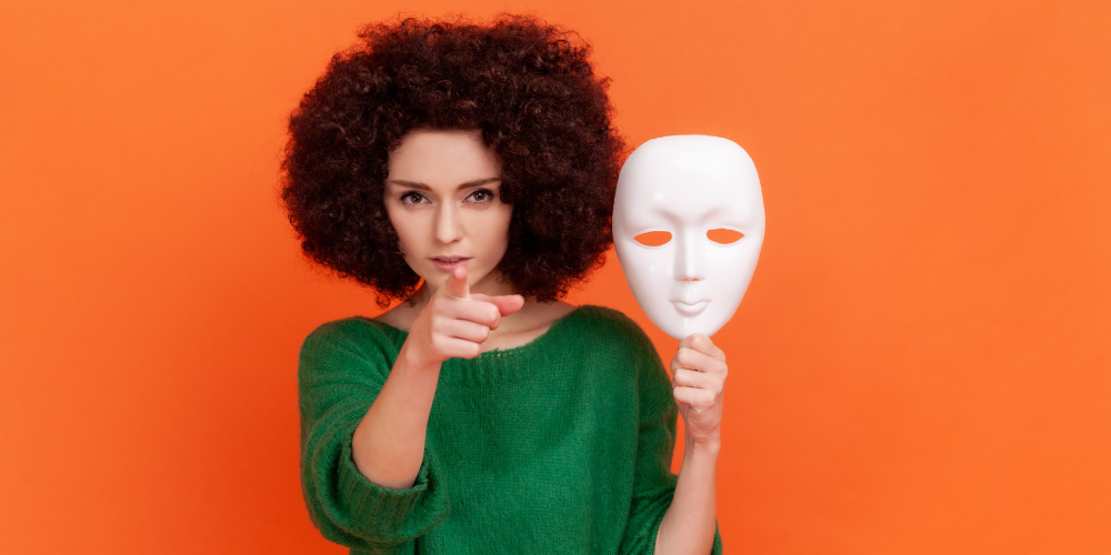 A woman holding a white mask and pointing forward with one finger symbolizing impostor syndrome
