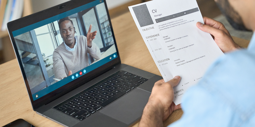 The image displays a person holding a printed CV while having a video call with a professional who appears to be discussing the CV or conducting an interview. The candidate is focused on the paper, and the interviewer is gesturing, likely explaining or asking a question. The setting suggests a remote job interview or consultation process.
