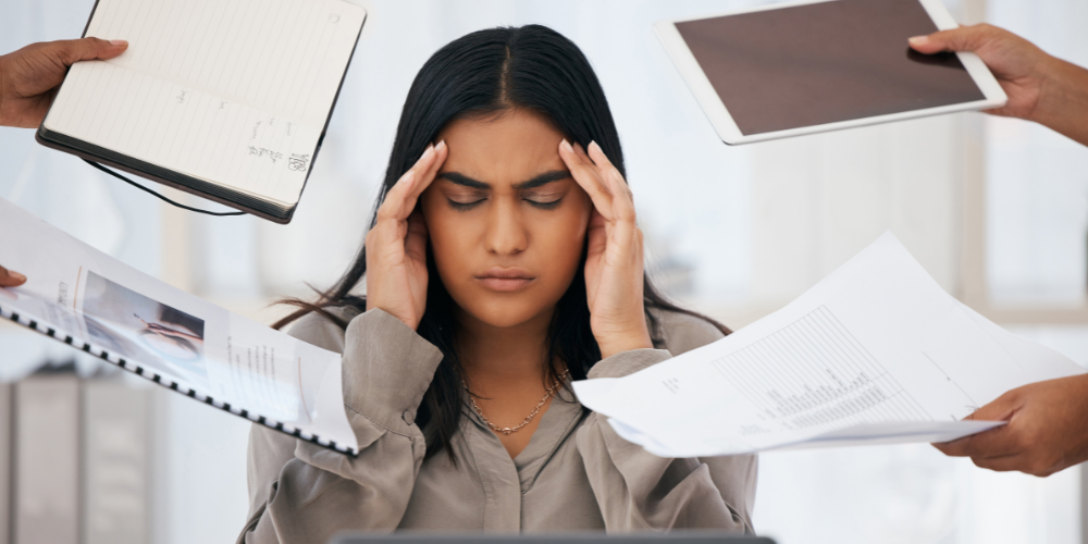 A stressed woman at work with her eyes closed and temples pressed by her fingers, surrounded by hands offering her multiple documents and a tablet, symbolizing overwhelming demands and the need for stress management.
