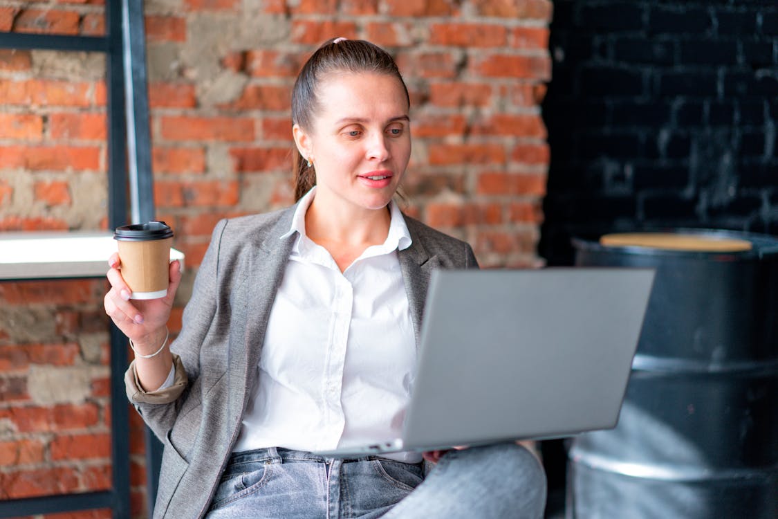 A woman is seated, holding a takeaway coffee cup in one hand and interacting with a laptop with the other. She's dressed in a casual business style, with a grey blazer over a white shirt and light grey jeans. The setting features an exposed brick wall with a window, giving the space an urban, modern feel.
