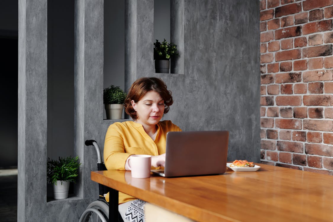 A woman is seated at a wooden table, focused on her laptop. She is wearing a yellow cardigan and is seated in a wheelchair. On the table, there is a pink mug and a plate with a light snack. Behind her is a wall with a mix of gray concrete and exposed brick textures, adorned with small potted plants, creating a cozy and stylish atmosphere.