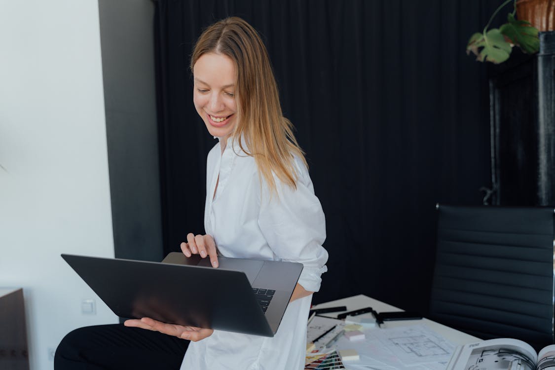 A woman in a white shirt is holding an open laptop with one hand working remote from home