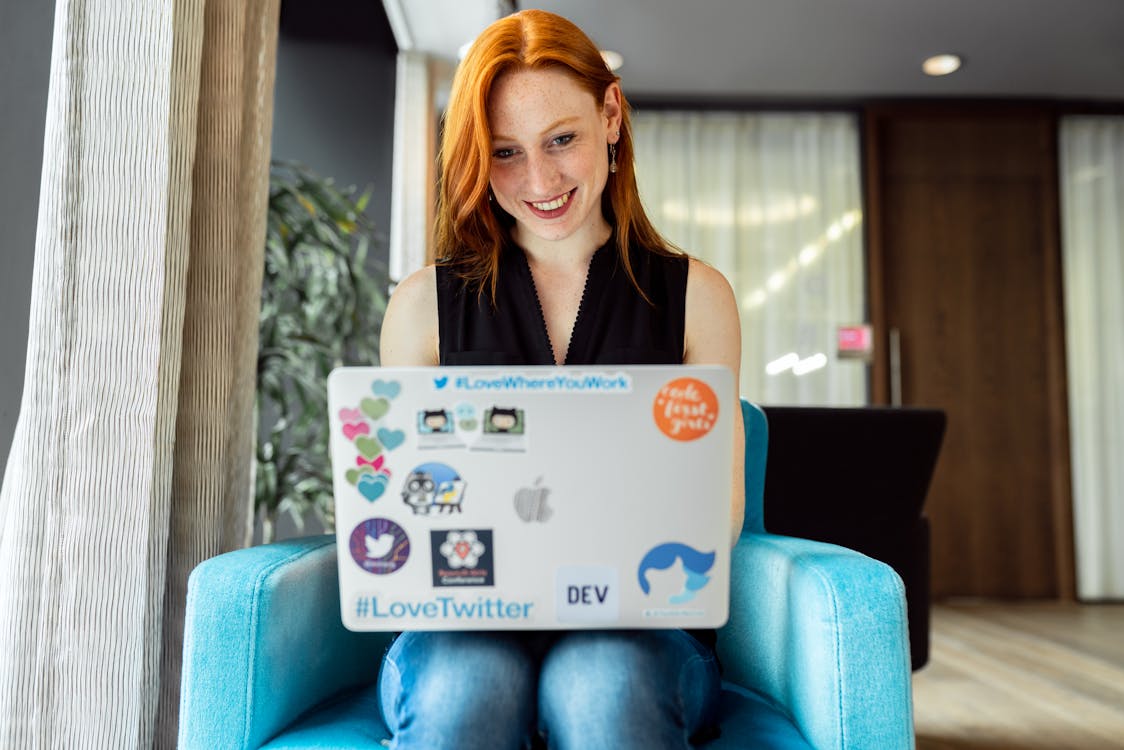 A smiling woman with red hair, wearing a sleeveless black top, is sitting in a blue armchair. She is using a laptop covered with various stickers, indicating a personalized touch. The setting appears to be a casual indoor environment, possibly a modern office lounge or coworking space.