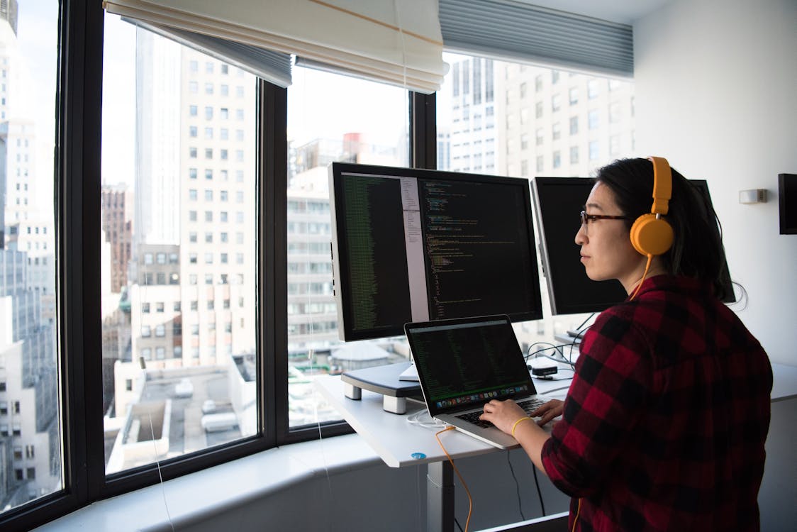 A person with glasses and wearing headphones is focused on coding on a laptop that is placed on a stand, with an additional monitor mounted above the laptop. They are wearing a red and black checkered shirt. The workstation is beside a window that offers a view of high-rise buildings, suggesting an urban office setting.
