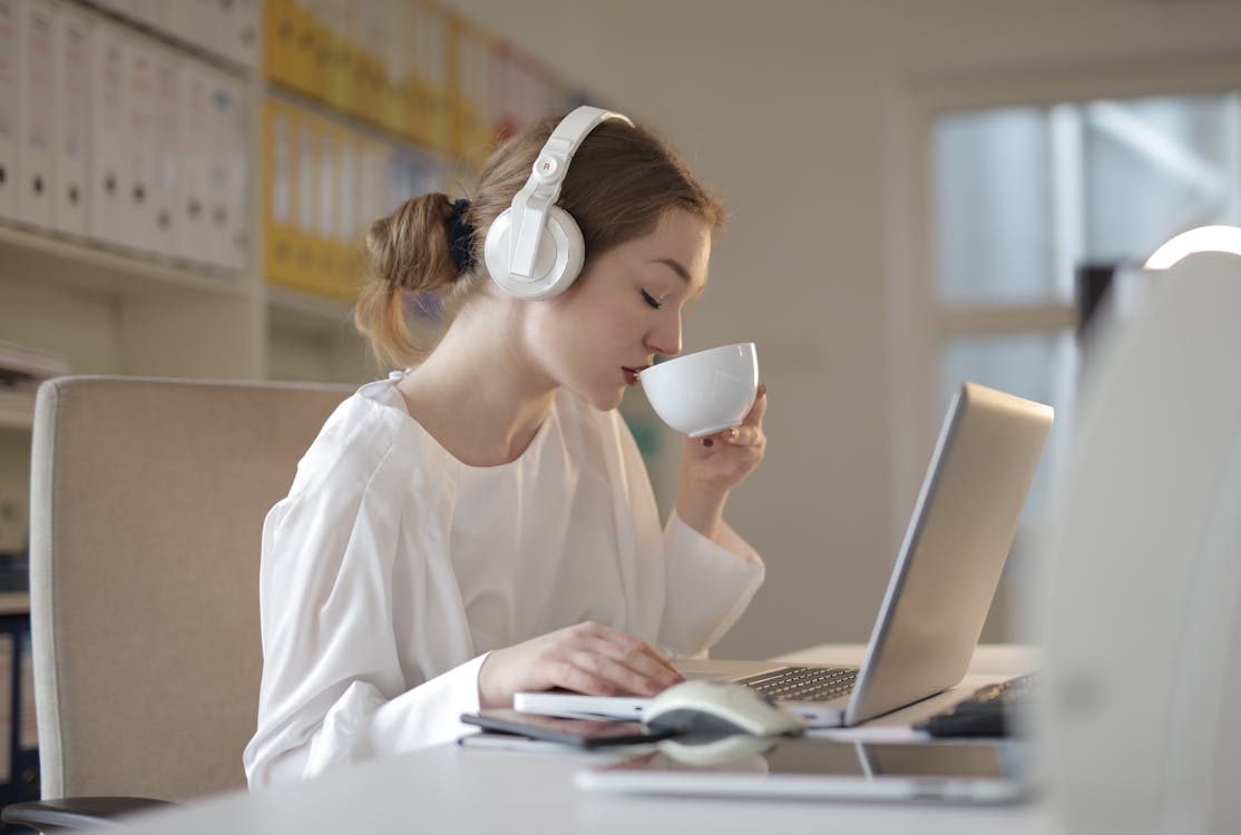 A woman with her hair in a bun, wearing a white blouse and over-ear headphones, is drinking from a white cup while working on a laptop in an office environment. There are file organizers in the background, and the setting appears to be well-lit with natural light.