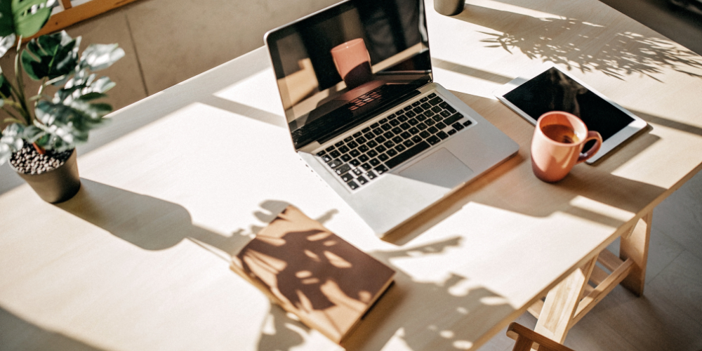 A cozy home office setup with natural sunlight casting shadows on a wooden desk, featuring an open laptop, a potted plant, a digital tablet, and a mug.