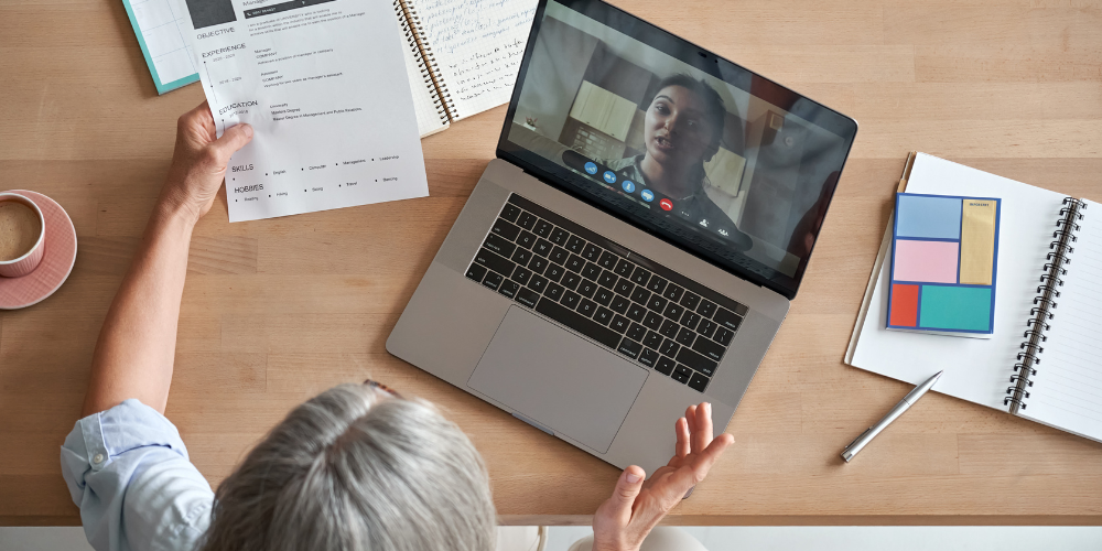 Overhead view of a person in a video call interview, with a resume in hand, and a notepad with sticky notes beside a laptop on a wooden desk.