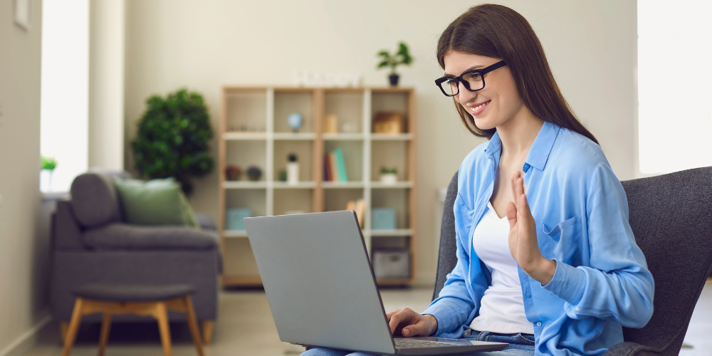 The image shows a cheerful woman sitting on a gray chair at home, greeting someone during a video call on her laptop. She's wearing glasses, a white shirt, and a blue shirt on top. The room is well-lit and has a modern, minimalistic design, with a green plant and a neat bookshelf in the background, indicating a clean and organized living space.
