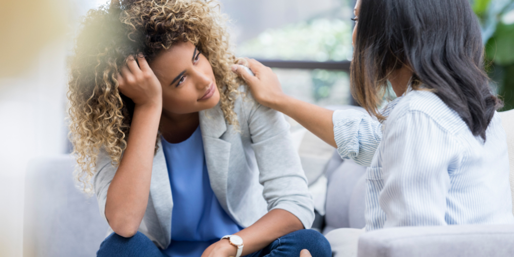 A compassionate colleague is comforting a distressed woman holding her head, demonstrating the importance of peer support for mental well-being in the workplace.