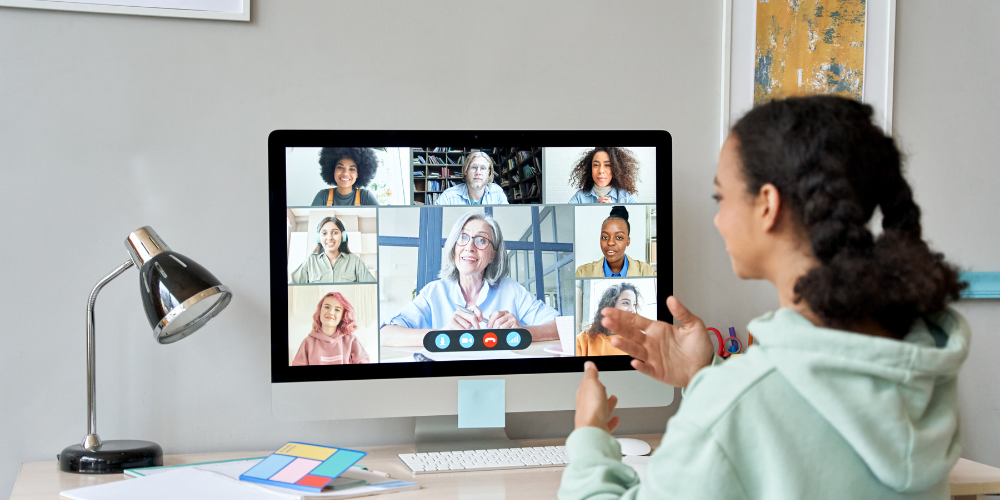 A woman engaging in a group video conference call on a desktop computer, with multiple participants on the screen, in a well-lit home office environment.