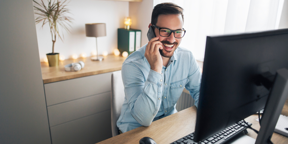 A cheerful man with glasses talking on a smartphone while working at his desk with a computer monitor, in a well-decorated home office with warm lighting.
