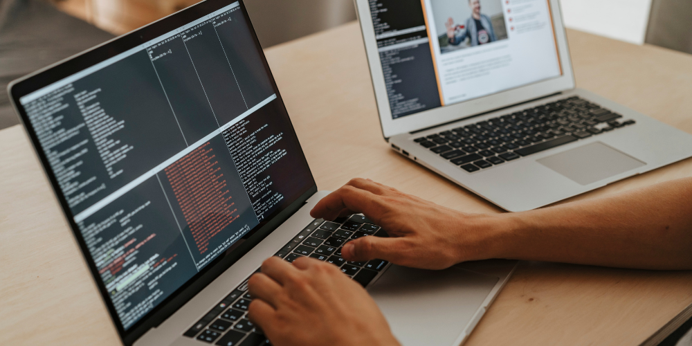 Close-up of a person's hands typing on a laptop with a second monitor displaying code in an integrated development environment, indicating a programming or software development task.