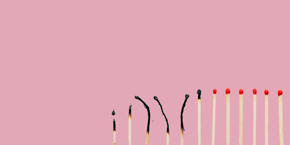 This image features a series of matches against a pink background. On the left, the matches appear burnt out with varying degrees of burn, symbolizing stages of burnout. To the right, the matches are unburnt, representing potential or energy yet to be expended. The visual metaphor illustrates the concept of burnout, with some matches completely spent, and others still intact, perhaps warning against the dangers of overextending oneself.