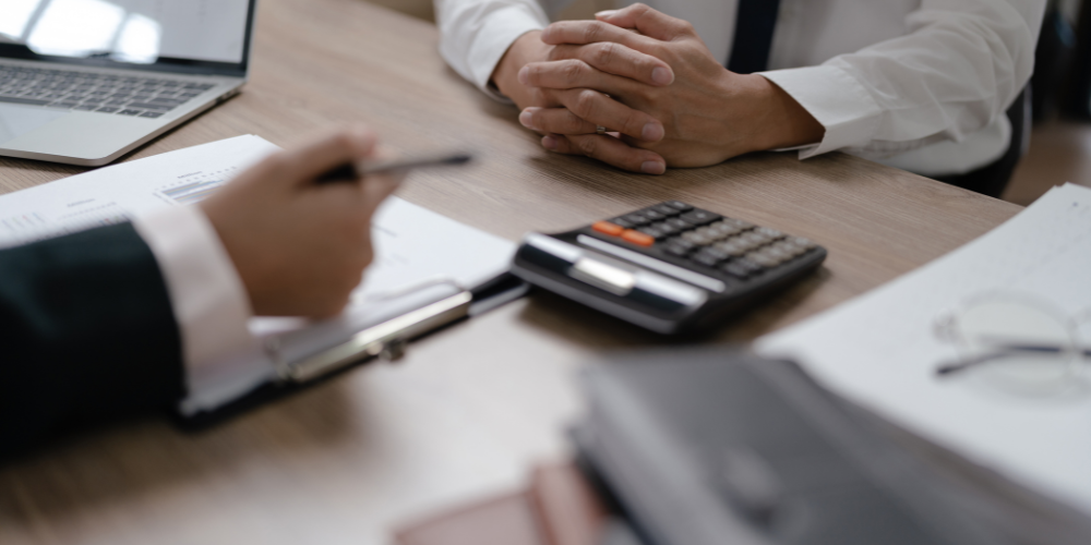 A close-up view of a professional setting where two individuals are engaged in a discussion with documents, a calculator, and a laptop on the table, suggesting a the calculation of the cost of employee turnover