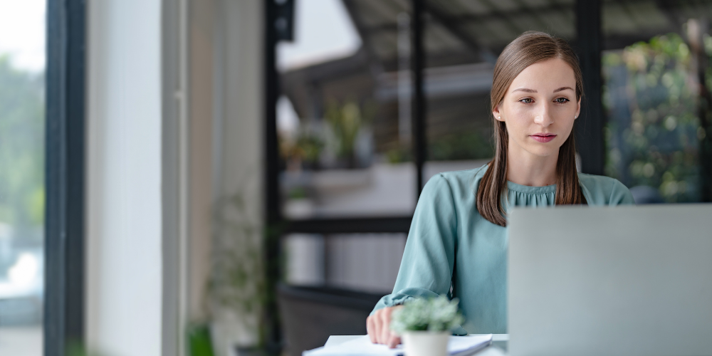 A focused woman working on a laptop in a well-lit office or home space with a blurred background, suggesting a professional or remote work environment.
