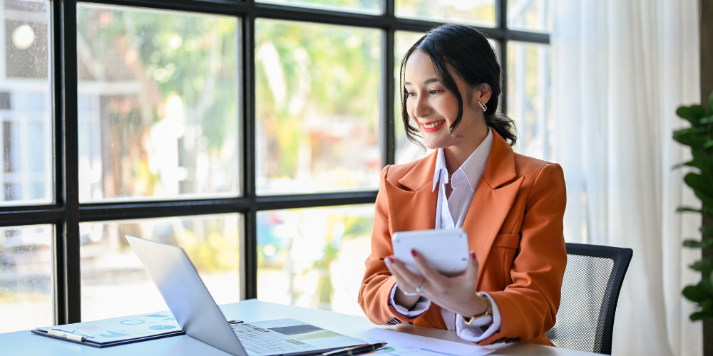 A woman in a stylish orange blazer is seated at a desk, smiling as she looks at her calculator, with a laptop and documents in front of her, suggesting she's happy after learning how to improve the employee turnover rate