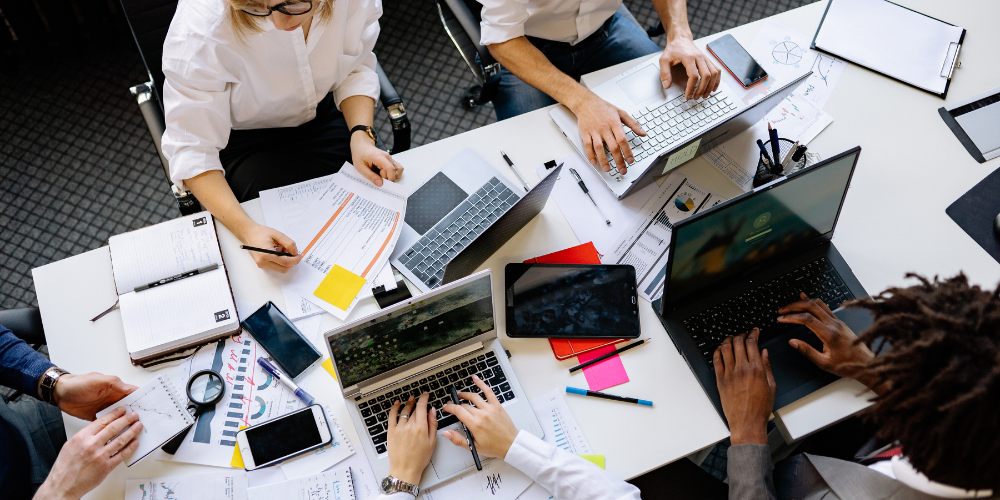 Overhead view of a dynamic brainstorming session with a diverse group of professionals engaged in discussion and working on laptops amidst a spread of documents and digital devices on a conference table.