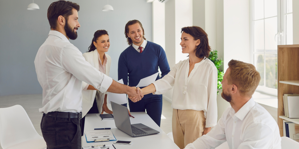 Two professionals shaking hands in a bright office setting with three colleagues looking on, embodying a collaborative work environment.