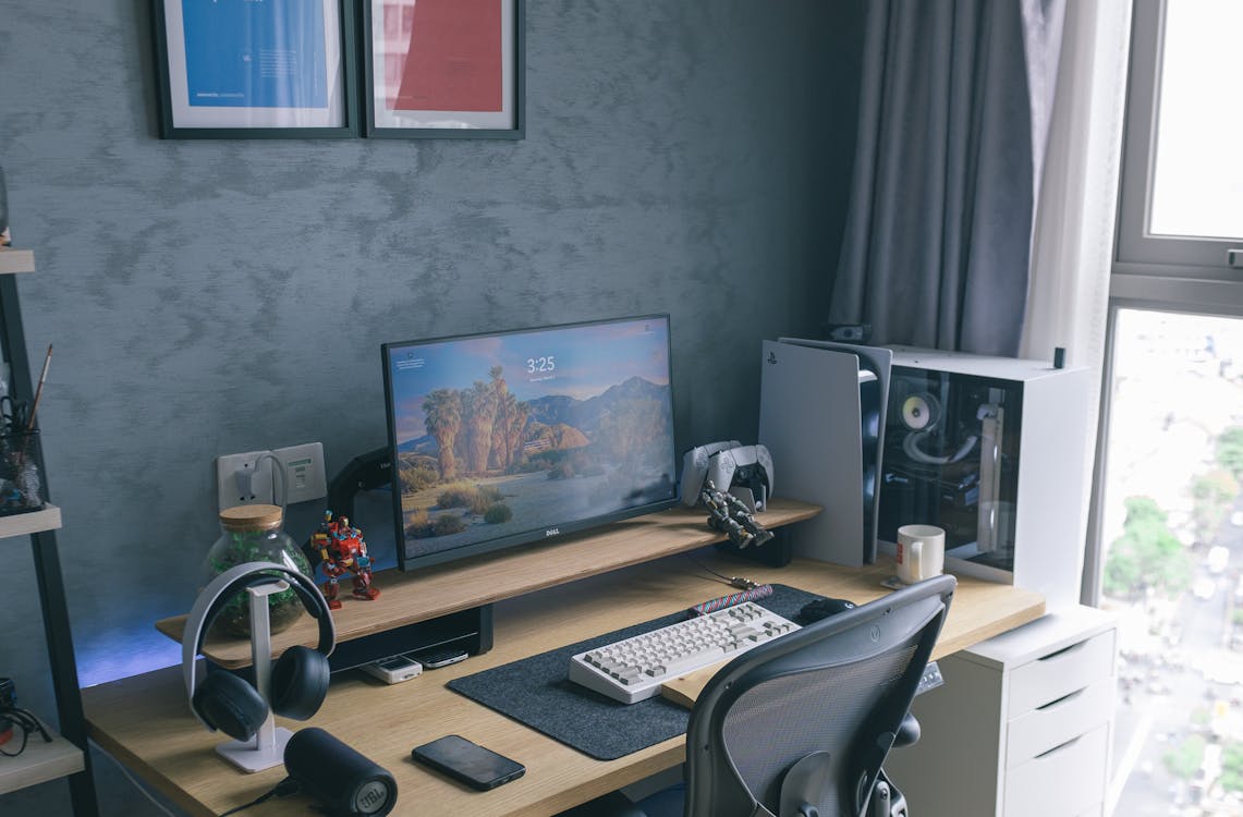 A well-organized work desk with a computer monitor, mechanical keyboard, and headphones stand, flanked by action figures and a desktop PC on the right. There are framed artworks on the wall, a bookshelf with various items, and a window with grey curtains, providing natural light.
