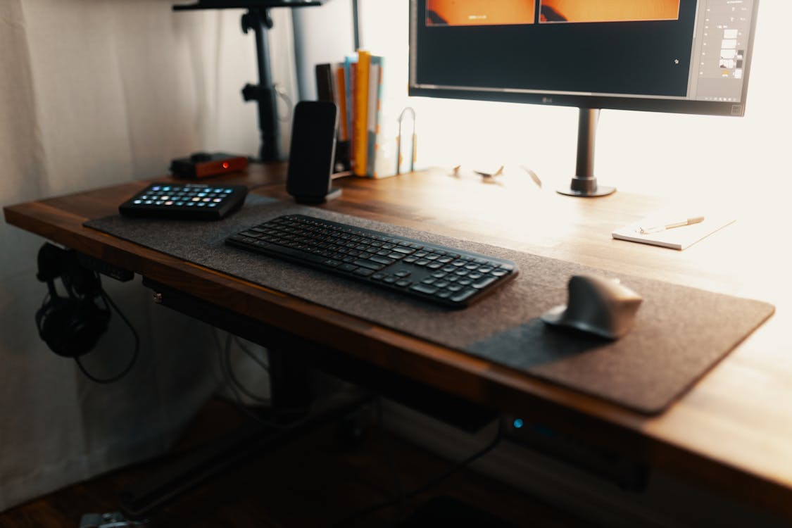 A home office setup with a wooden desk featuring a computer monitor, a keyboard on a desk mat, a smartphone, a tablet, headphones hanging on the side, and some books and stationery items in the background, illuminated by natural light.