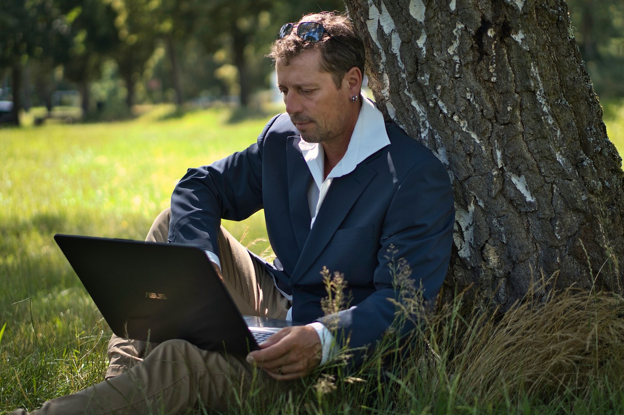 A man in a suit working on a laptop outdoors, sitting at the base of a tree.