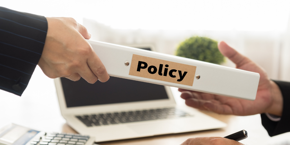 Two hands in business attire passing a binder labeled "Policy" across a desk with a laptop and other office items in the background.