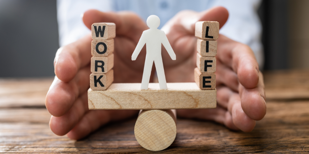 A person's hands holding a wooden balance toy with a figure standing in the middle; one side is labeled "WORK" and the other "LIFE" to represent work-life balance.