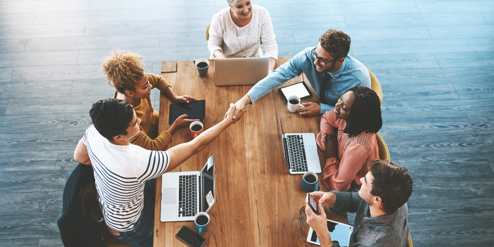 Overhead view of a diverse group of professionals engaging in a friendly meeting around a wooden table, with laptops, smartphones, and coffee cups, showcasing teamwork and collaboration in a modern workplace