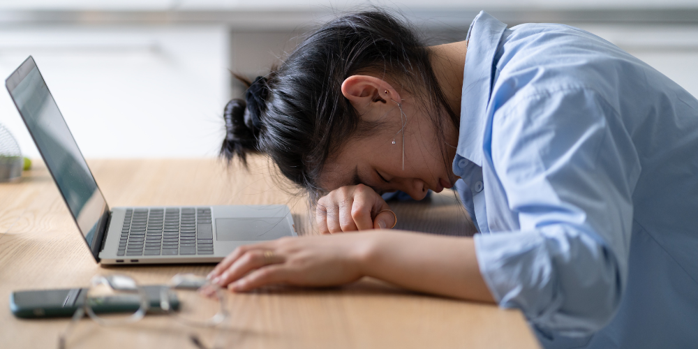 This image shows an individual resting their head on a desk in front of an open laptop, suggesting a state of exhaustion or burnout. Nearby items include a pair of glasses and a smartphone, indicating that work and communication tools are at hand but the person is perhaps too fatigued or overwhelmed to use them. The setting seems to be a personal workspace or home office.