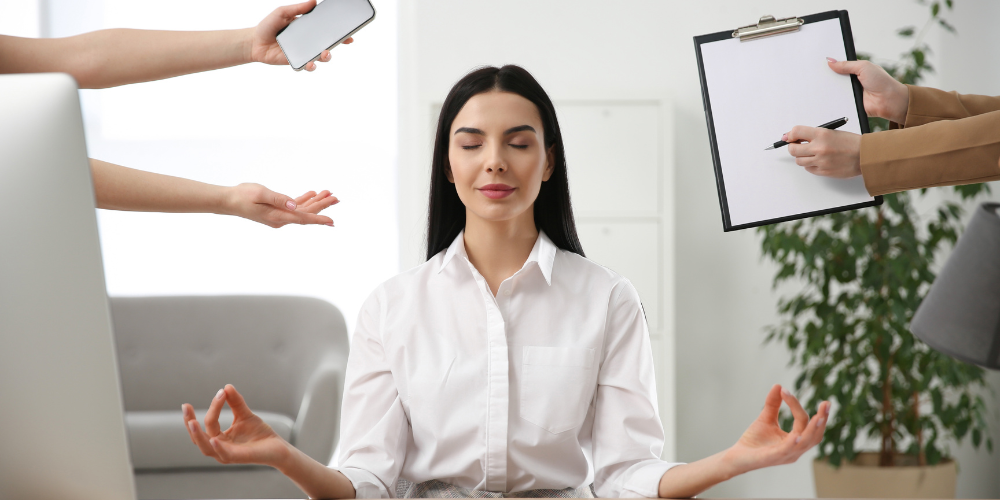 A woman meditates at her desk, ignoring surrounding hands offering a phone and clipboard, symbolizing tranquility amidst workplace demands.