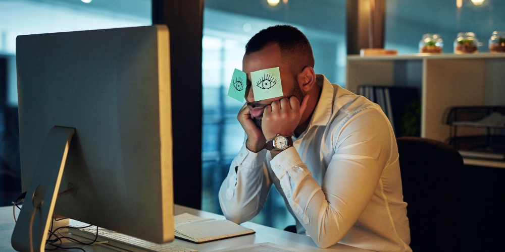 The image shows a person sitting at a desk, facing a large desktop computer monitor. They are holding a sticky note with drawn eyes over their face, which creates the humorous illusion that they are awake and alert. This scene often represents the idea of someone trying to appear awake or attentive, potentially during a tedious task or late hours, and could imply fatigue or a desire to mask true feelings of tiredness or boredom. The environment suggests a modern office or home workspace.