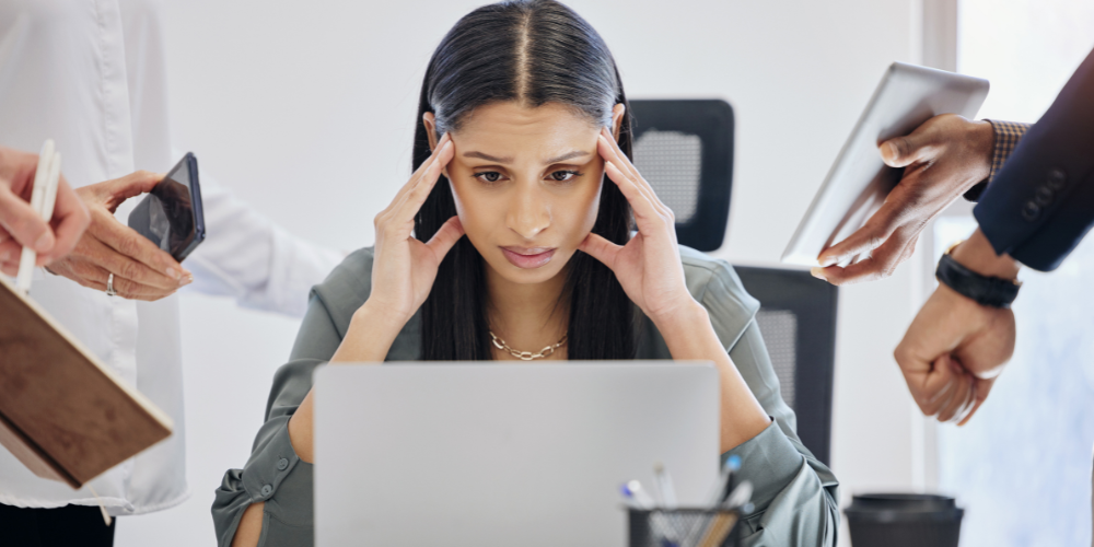 a woman sitting at a desk looking at a laptop screen with a stressed or overwhelmed expression. Surrounding her are hands from different people offering her various items such as a phone, a tablet, and a notebook, which suggests that she is being given multiple tasks or information at the same time.