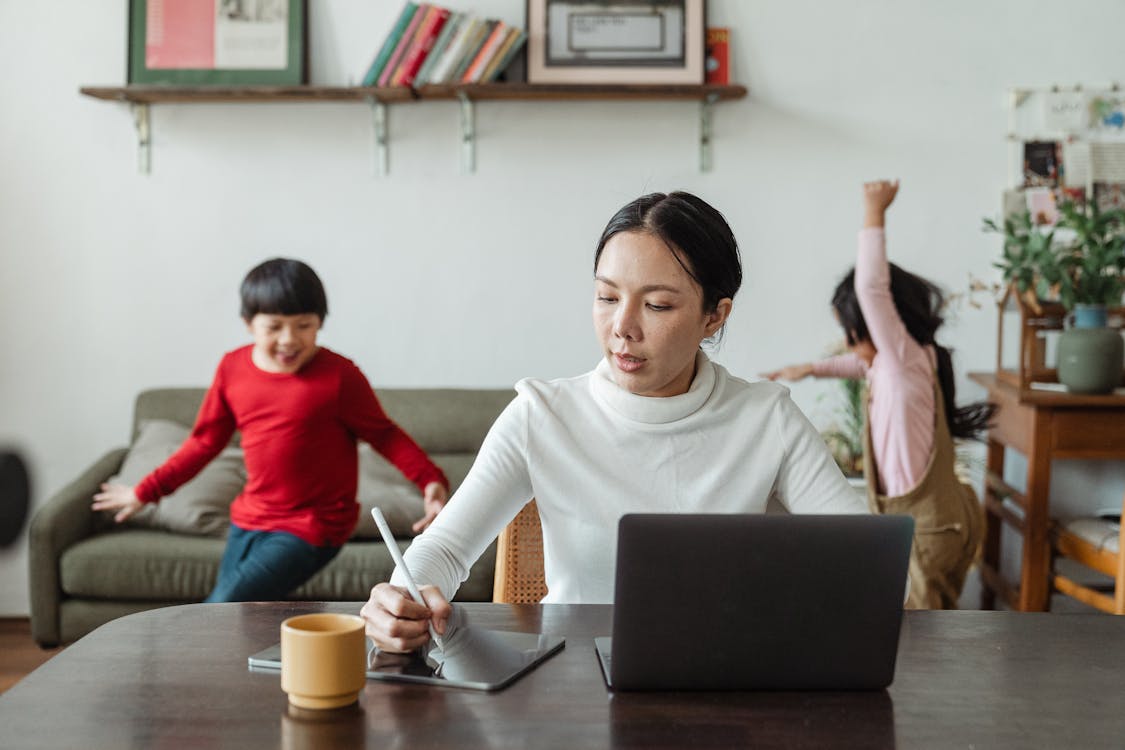 A focused woman working on a laptop and using a stylus on a digital tablet at a home office desk, with two children playing in the background, one in a red shirt and the other in a pink top and beige pants. There are shelves with books and plants in the room.