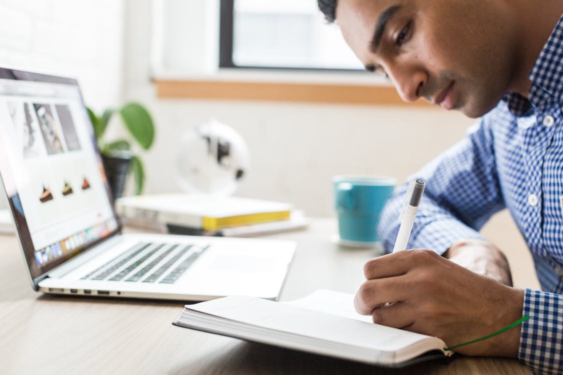 A focused individual in a blue checked shirt writing in a notebook, with a laptop open in front of him displaying images, a blue mug, and a plant in the background, indicating a casual work or study environment.