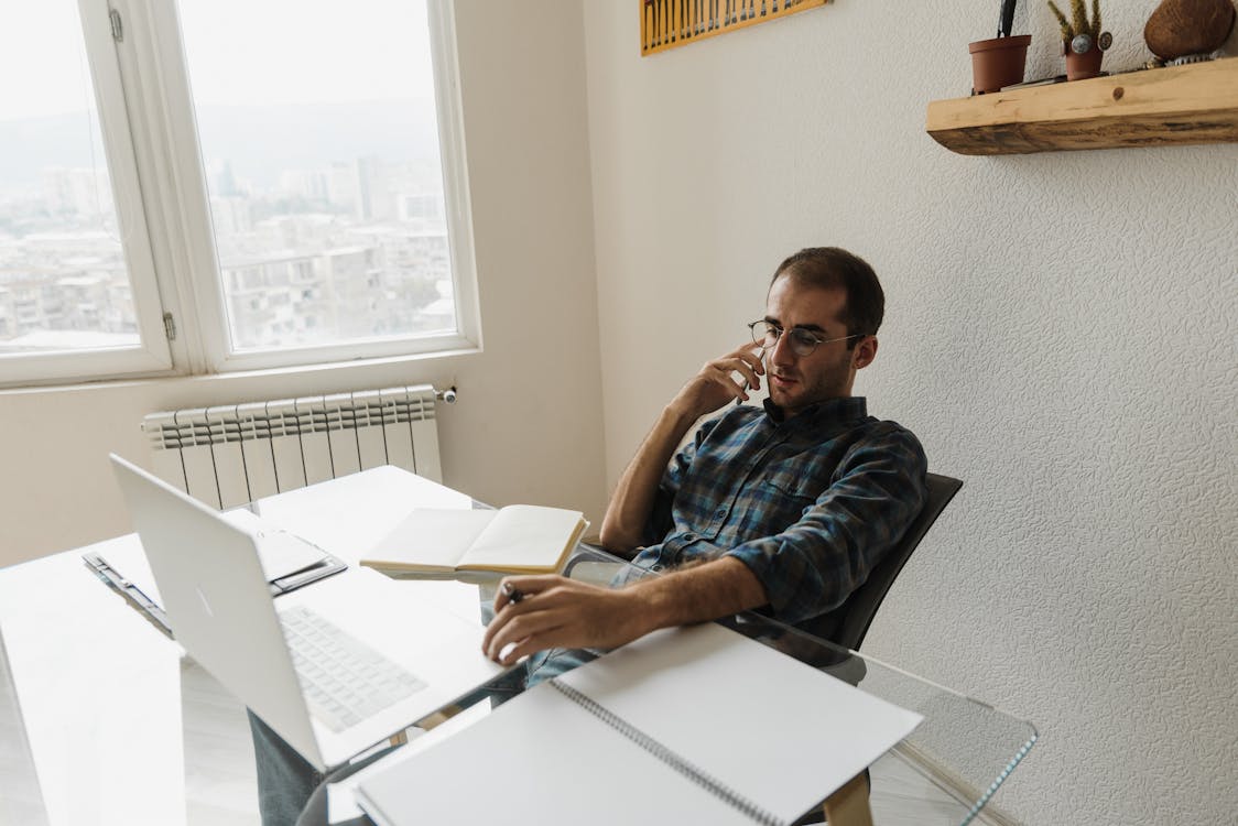 A man with glasses, wearing a plaid shirt, speaking on a smartphone with a pensive expression, seated at a desk with a laptop, notebooks, and a book, in a well-lit room with a window offering a city view and a shelf with plants above.