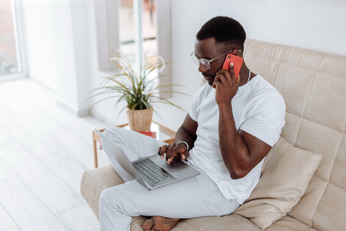A man in white clothing sitting on a beige couch, talking on a red phone while looking at a laptop placed on his lap, with a potted plant in the background near a bright window.