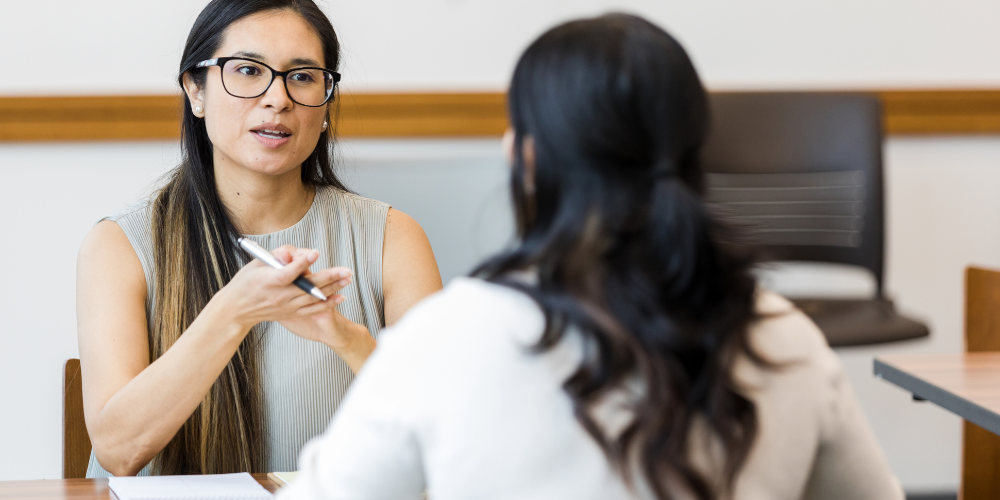 Two women engaged in a professional conversation, with one facing the cam-velascoera holding a pen and gesturing, wearing glasses and a sleeveless top, and the other with her back to the cam-velascoera, in a business casual setting.