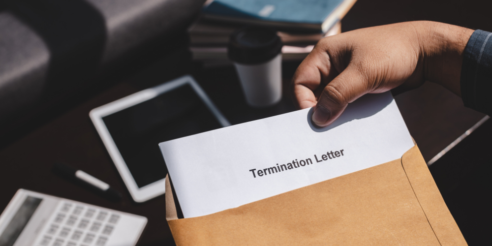 A person's hand pulling a document labeled 'Termination Letter' out of a brown envelope, with office items like a calculator, tablet, and coffee cup in the background on a dark desk surface.