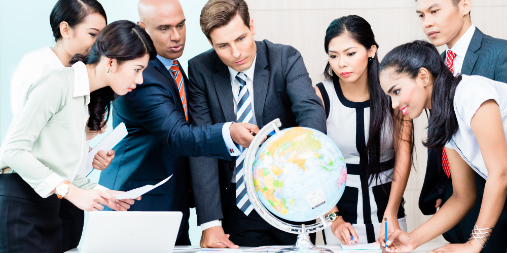 A group of professionals gathered around a globe, pointing and looking at various locations, indicating a discussion on global affairs or international business. They appear engaged and focused, with documents and a laptop indicating a working session or meeting.