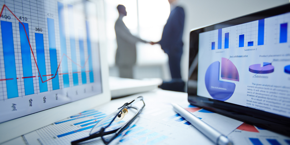 Foreground shows a close-up of business analytics displayed on a computer screen with graphs and charts, with eyeglasses and a pen suggesting a work environment. In the blurred background, two individuals appear to be shaking hands, implying a successful business agreement or partnership.