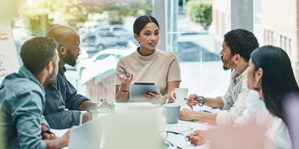 A diverse group of professionals is engaged in a meeting around a table. A woman appears to be leading the discussion, holding a tablet and addressing her colleagues. The setting suggests a collaborative, modern office environment.