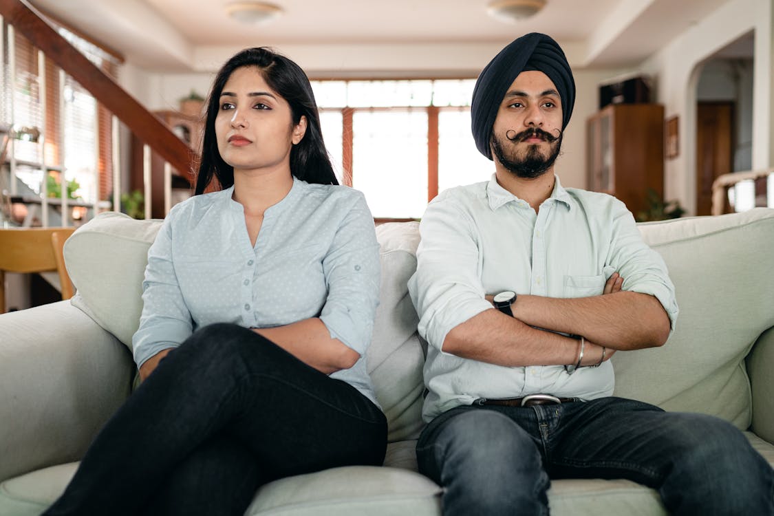 A man in a turban and a woman sitting side by side with serious expressions, arms crossed, on a sofa in a home setting.